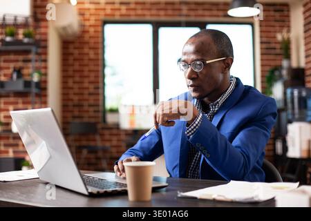 African american male analyst seated at desk with pen in hand, analyzing online financial documents on his laptop in startup office. Focused black man using digital device to research strategy ideas. Stock Photo