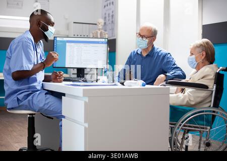 Attentive male healthcare worker uses computer to guide elderly woman in wheelchair with customized health plan. African american nurse and caucasian old couple wearing face mask in medical office. Stock Photo