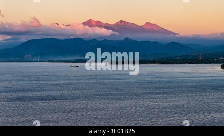 Active volcano Mount Rinjani glowing pink at sunset over the sea in Lombok, Indonesia Stock Photo