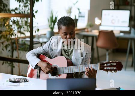 Young Adult Black Woman Playing Acoustic Guitar While Sitting at Desk Indoors Stock Photo