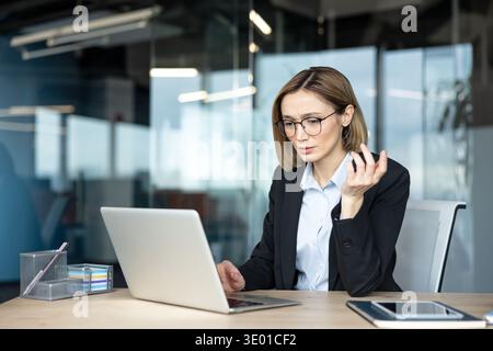 Businesswoman thinking critically while working on a laptop, facing a complex problem or project in a modern office environment, contemplating solutions with a focused and frustrated expression Stock Photo