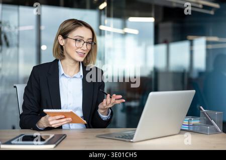 Professional businesswoman engaging in a remote video conference, taking notes in a notebook, and using a laptop in a modern office, representing virtual communication and distance learning concepts Stock Photo
