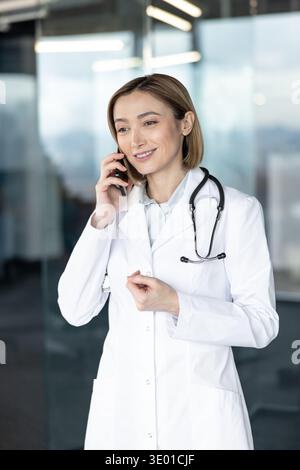 Female doctor in white coat with stethoscope on shoulders smiling while having a phone conversation, representing healthcare professional communication and telemedicine services Stock Photo