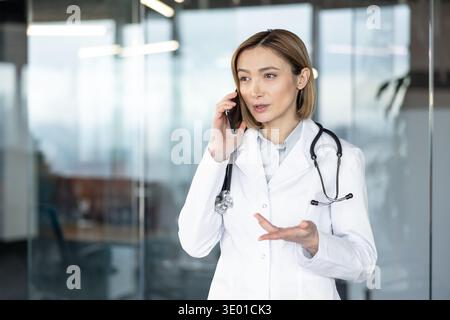 Female doctor in white coat with stethoscope in a modern clinic, speaking on a smartphone during a focused teleconsultation, explaining care and offering confident advice Stock Photo