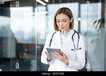 Female doctor in a white lab coat and stethoscope is carefully reviewing medical data on a digital tablet, standing in a contemporary office setting, representing modern healthcare technology Stock Photo