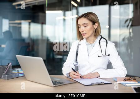 Doctor in white coat and stethoscope using laptop for medical research while writing on clipboard paper, working on healthcare documentation in a modern office Stock Photo