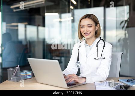 Female doctor working on a laptop, smiling at the camera, wearing a lab coat and stethoscope in a modern office, representing healthcare innovation and professional medical consultation Stock Photo