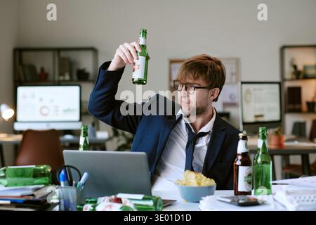 Young Caucasian Man Sitting at Desk Drinking Beer While Working on Laptop Stock Photo
