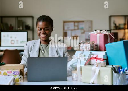 Young Adult Black Woman Smiling While Working on Laptop Surrounded by Gifts Stock Photo