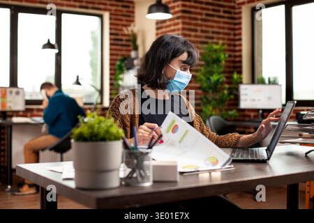 Female analyst sits at office desk with financial paperwork and checks company research data on laptop. Young woman with face mask using digital device and documents to review business analytics. Stock Photo