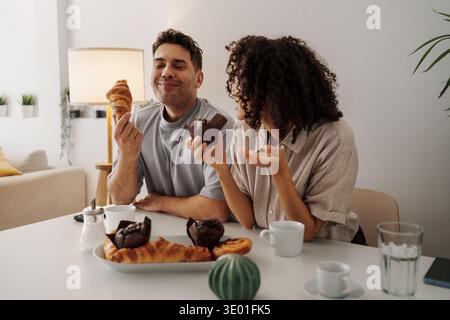 Joyful Breakfast Chat. Cheerful Duo Sharing Flaky Croissant And Steaming Coffee In Cozy Setting Stock Photo