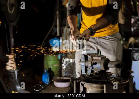 Focused worker man in workshop at night, grinding metal with tool. Sparks fly from industrial machine, showcasing skill and precision in dark setting Stock Photo
