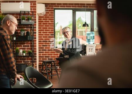 Smiling retired woman psychologist welcoming her clients in office, ready for group therapy session. Selective focus on elderly female psychiatrist with clipboard and gesturing to group of people. Stock Photo