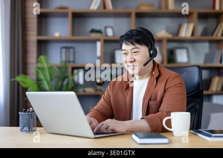 Smiling asian man in a home office wearing a headset and typing on a laptop while providing customer support or joining a virtual meeting, focused and professional in a casual workspace Stock Photo