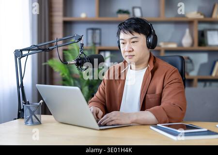 Asian man podcast host wearing headphones and speaking into a studio microphone while typing on a laptop in a modern home office, focused on recording and content creation Stock Photo