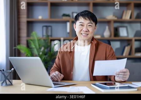 Happy asian man smiling at camera while sitting at his home office desk, holding a stack of papers and working on his laptop, embodying successful remote work and productivity Stock Photo