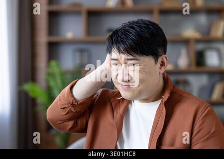 Thoughtful asian man experiencing stress and confusion, holding hand to his head while working in a modern home office setting, contemplating or dealing with a difficult situation Stock Photo
