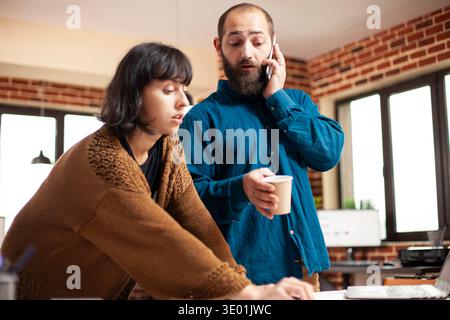 Male employee speaking on phone with client and reviewing business project presentation with coworker. Businessman talks on mobile device as female colleague helps review company research documents. Stock Photo