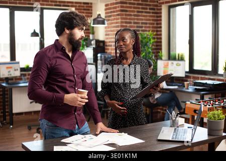 Caucasian businessman holds coffee cup and listens to black woman with pregnancy belly share startup strategy on clipboard. Multiethnic coworkers collaborating on company project in brick wall office. Stock Photo