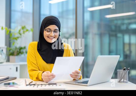 Muslim businesswoman wearing a hijab and glasses, smiling while reviewing documents at her office desk with a laptop, reflecting work satisfaction and professional success Stock Photo