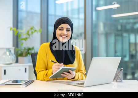 Muslim businesswoman in a hijab smiling and taking notes in a planner during an online meeting on her laptop in a modern office, focused and confident at her desk Stock Photo