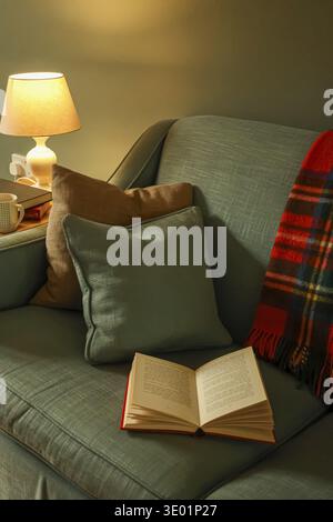 Open book resting on a sofa with pillows and a plaid blanket, lit by a warm table lamp creating a cozy atmosphere Stock Photo