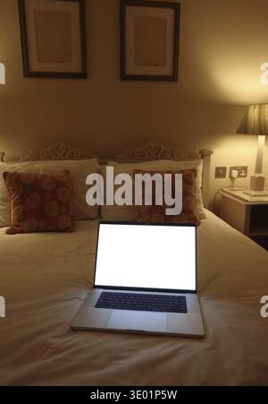 Laptop with blank white screen displaying mockup content, resting on a bed in a hotel room or bedroom. Mockup copyspace Stock Photo