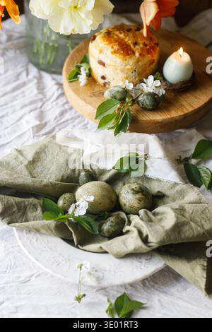 Happy Easter! Stylish eggs on napkin on plate, easter cake on stand, spring flowers and candle. Easter celebration table setting, holiday still life. Stock Photo