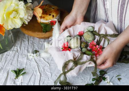 Easter celebration. Hands holding stylish green easter eggs on napkin in plate on background of easter bread and cherry blossoms on table. Atmospheric Stock Photo