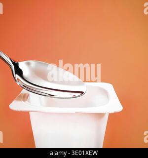 Yogurt cup and silver spoon on orange background, white plastic container with yoghurt cream, fresh dairy product for healthy diet and nutrition balan Stock Photo