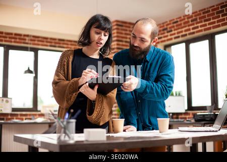 Caucasian male and female colleagues looking at company research data on clipboard. Bearded man and young woman standing at office desk and reviewing financial paperwork in brick wall room. Stock Photo