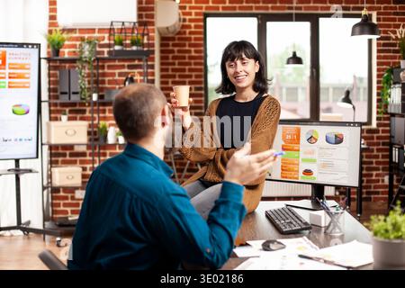 Smiling businesswoman with coffee, sitting on office desk and discussing with male coworker in brick wall room. Caucasian colleagues taking a break and having conversation in startup office. Stock Photo