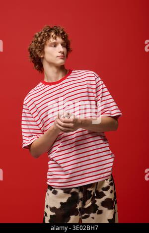 A stylish young man with curly hair stands confidently in a red studio, wearing stripes. Stock Photo