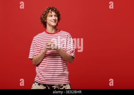 A fashionable young man stands confidently in a red studio, smiling with hands clasped. Stock Photo