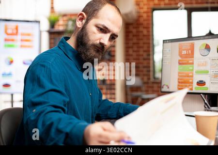 Closeup of bearded man seated at desk, focused on reviewing market analysis documents. Male manager looking at financial paperwork as computer displays graphs and analytics tools in brick wall office. Stock Photo