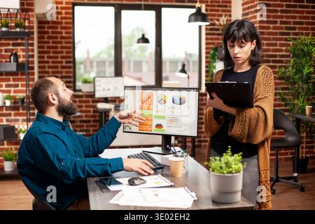 Young woman stands in front of colleague, holding clipboard and confidently explaining project updates. Supportive bearded man listening to female coworker, ready to collaborate on company research. Stock Photo