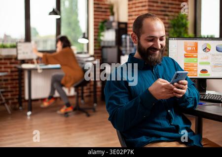 Happy businessman sits at shared brick wall workspace, texting on mobile device. Male employee checking social media on cellphone as computer screen displays business analytics in startup office. Stock Photo