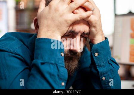 Closeup of male analyst holding his head in hands, having exhausted expression from reviewing financial analysis. Bearded businessman with eyes closed, feeling tired and overwhelmed in startup office. Stock Photo