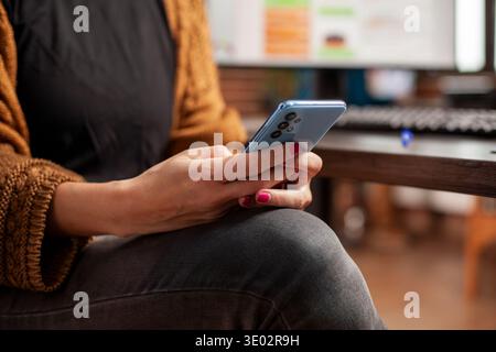 Selective focus on hands of caucasian woman texting on smartphone while seated at office desk. Closeup of female employee holding mobile device, browsing social media during working hours. Stock Photo