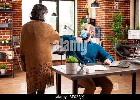 Young man and woman in business casual clothing greet each other with elbow bump at desk in brick wall room. Caucasian colleagues wearing face masks and ensuring pandemic safety in startup office. Stock Photo