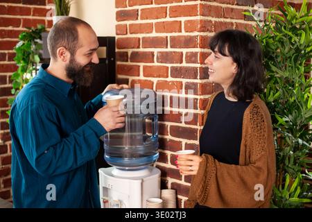 Smiling caucasian employees hold coffee cups, engaging in casual yet productive conversation in brick wall office. Businessman and woman having relaxed interaction with refreshment near water cooler. Stock Photo