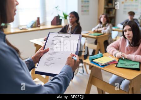 Hispanic teacher holding clipboard with test paper standing in classroom, multiethnic group of teenagers sitting at desks listening attentively during lesson, educational setting Stock Photo