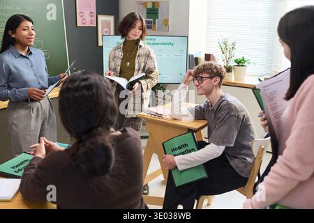 Group of multiethnic teenagers discussing health education in classroom, female teacher standing nearby guiding students, young male teenager holding health education folder, classmates listening Stock Photo