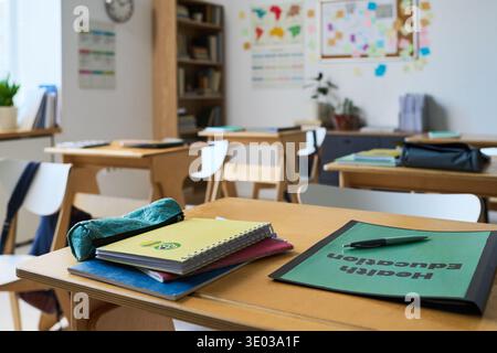 Empty classroom showing wooden desks with notebooks, folders labeled Health Education, pen and pencil case arranged on surface, bookshelves and educational posters visible in background Stock Photo