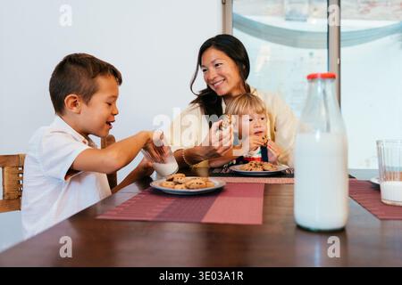 Asian family at home - asian mother and two sons sharing cookies and milk at home, smiling and bonding during cozy snack time together Stock Photo
