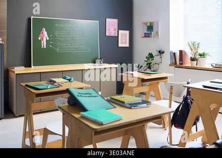Empty classroom featuring wooden desks and chairs arranged in rows, green chalkboard displaying human body diagram and health education notes, sunlight streaming through window blinds Stock Photo