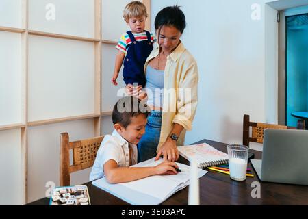 Asian family at home - asian mother helping older son with homework and drawing while holding toddler at home, supportive family learning moment Stock Photo