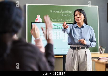 Young adult Hispanic woman standing in classroom pointing at student while teaching lesson, two students with raised hands sitting in foreground, green chalkboard and digital screen in background Stock Photo