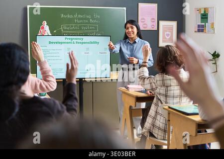 Young adult Hispanic woman teaching health education lesson about pregnancy and responsibility to group of teenagers in classroom, students raising hands to participate Stock Photo