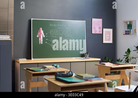 Empty classroom showing desks with books and supplies, green chalkboard displaying human body diagram and handwritten lesson topics about skeletal, muscular, nervous, reproductive systems Stock Photo